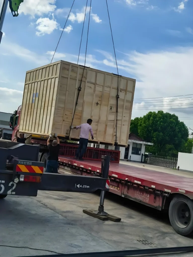 A large wooden export crate being lifted by a crane onto a red flatbed truck, with workers guiding the load, demonstrating safe handling of heavy industrial equipment during road transport.
