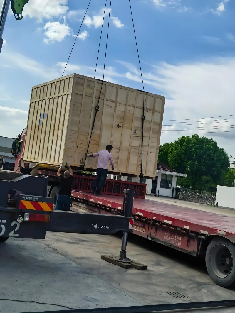 A large wooden export crate being lifted by a crane onto a red flatbed truck, with workers guiding the load, demonstrating safe handling of heavy industrial equipment during road transport.