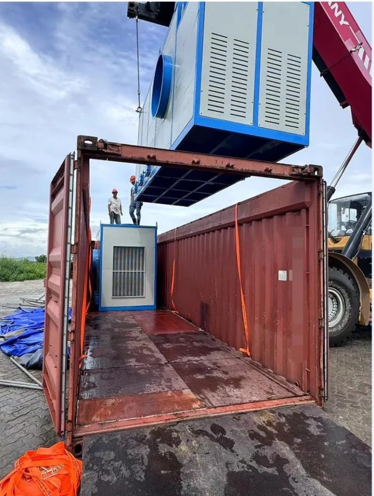 Workers guide a large blue industrial unit as it is lowered by crane into an open top container, demonstrating precise coordination required for safe oversized cargo loading.
