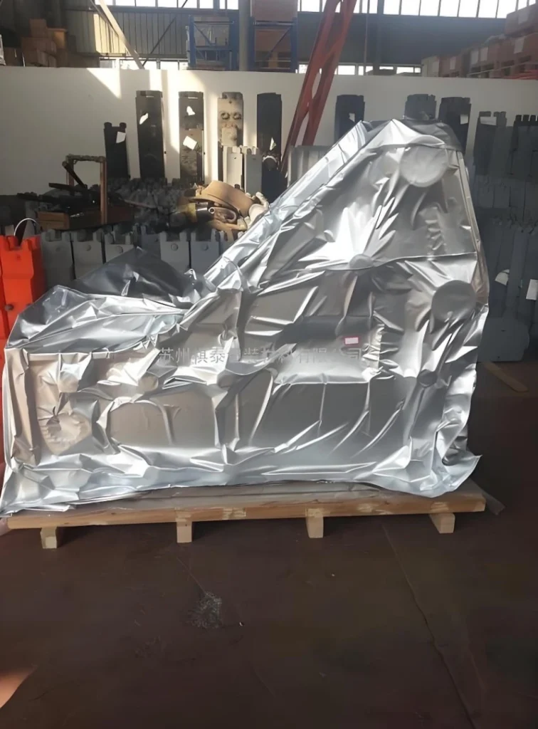 A uniquely shaped piece of industrial machinery fully encased in reflective aluminum foil vacuum packaging on a wooden pallet, showcasing customized barrier film solutions for complex equipment geometries.