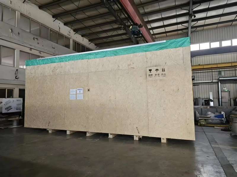 A large plywood export crate with handling symbols and green moisture barrier lining sits inside a warehouse, ready for shipment of heavy machinery or precision instruments.