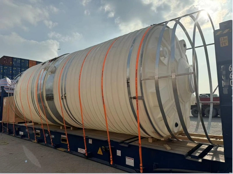 A large, white, ribbed cylindrical PE plastic storage tank is transported on a flatbed trailer, secured with multiple orange lashing straps and supported by a custom metal cradle to prevent deformation.