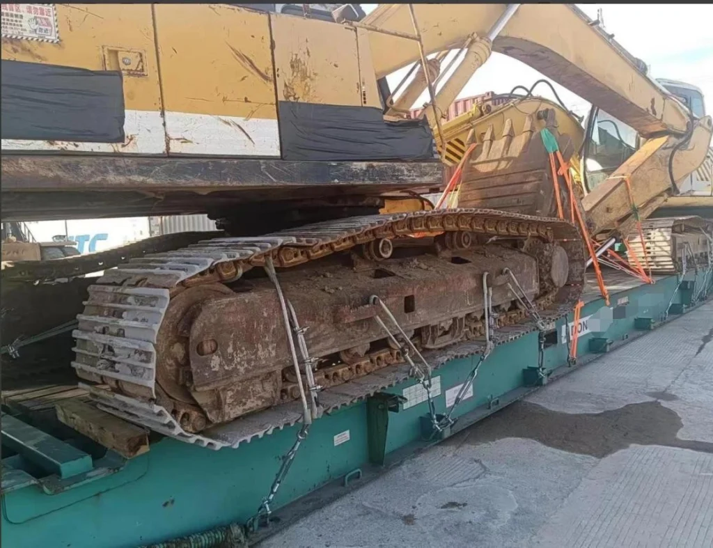 Close-up shot showing the muddy tracks of an excavator chained and strapped to a green flat rack base, highlighting specific lashing points and safety measures for heavy haulage.