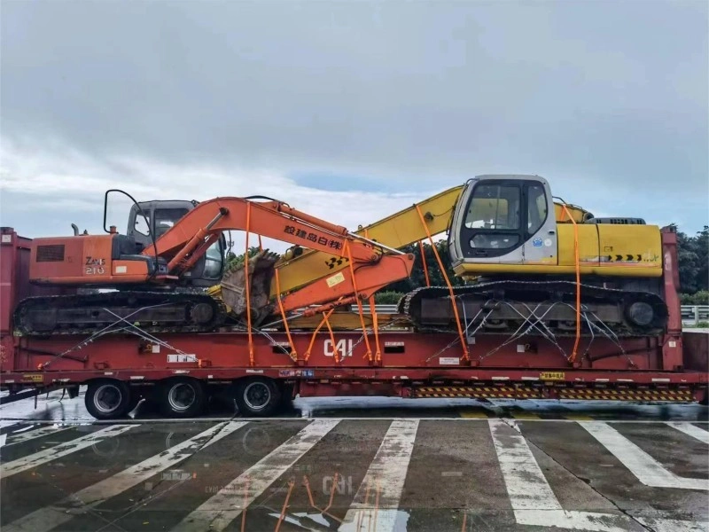 An orange ZAXS 210 and a yellow excavator are positioned side-by-side on a multi-axle red lowbed trailer, secured with cross-lashing straps, preparing for road transport under overcast skies near a paved crossing zone.