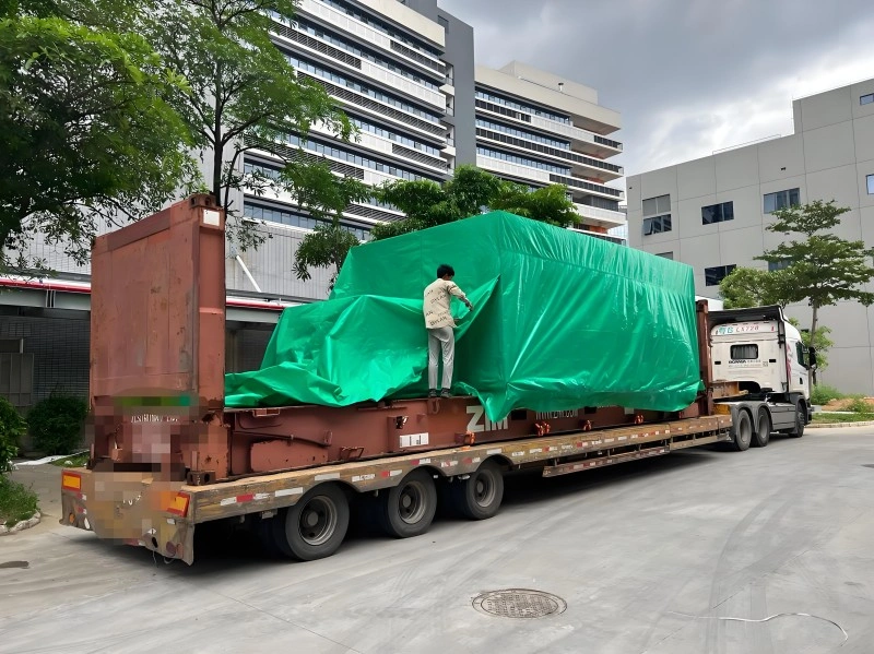 Two separate large industrial units wrapped in green protective covers and cross-lashed with orange straps on a single flat rack container, ready for export shipment at a logistics facility.