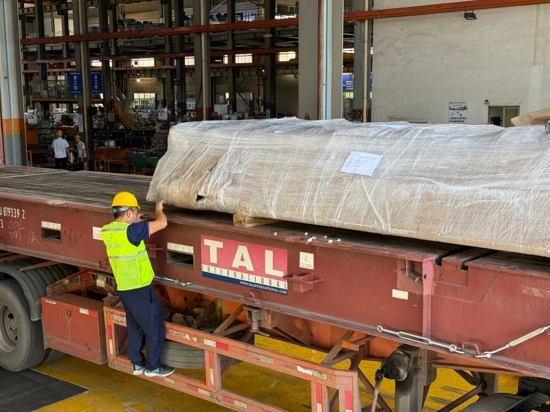 Worker in yellow hard hat and Bentlee safety vest conducting a final inspection of a large, plastic-wrapped industrial machine secured on a red TAL flatbed trailer in a warehouse