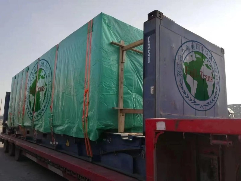 A side view of a flat rack container on a trailer, loaded with a large cargo unit wrapped in a green tarpaulin. The cargo is secured with orange straps and supported by wooden beams. The container end wall features a logo with a globe and the text "Full Trans".