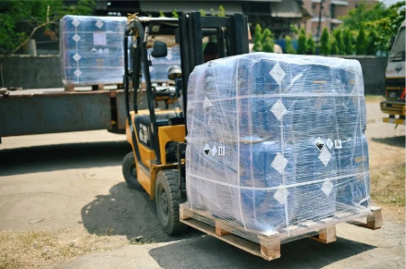 Yellow forklift lifting a palletized load wrapped in clear plastic film with diamond-shaped hazard labels, illustrating proper lifting technique to prevent bottom seal damage.