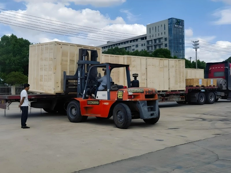 An orange forklift maneuvering a large wooden export crate onto a red flatbed truck, with workers supervising the operation, illustrating efficient and secure loading procedures for industrial shipments.