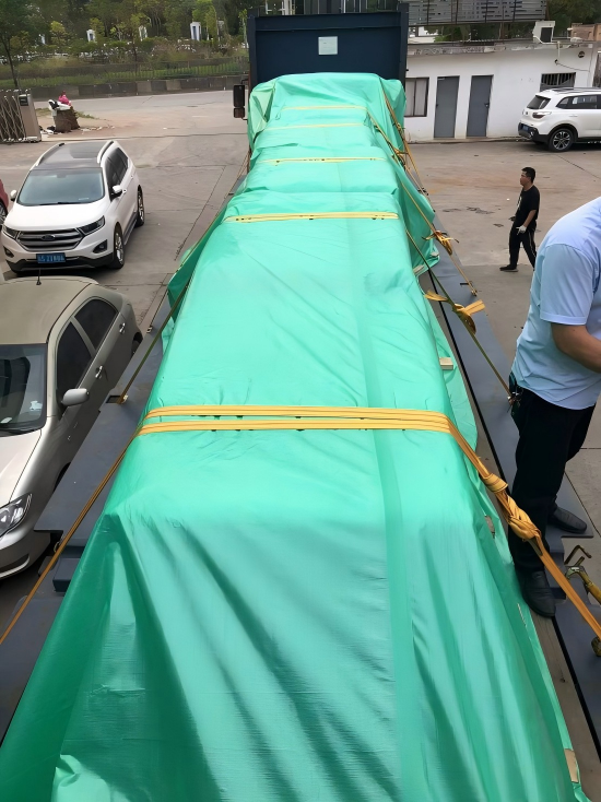 A large industrial mold covered with a green tarp and yellow straps, ready for road transport from Dongguan to the port