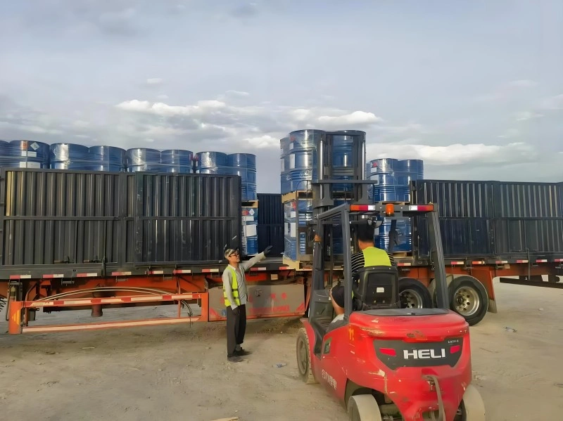 Workers loading blue chemical drums onto a truck trailer using a red HELI forklift in an outdoor yard