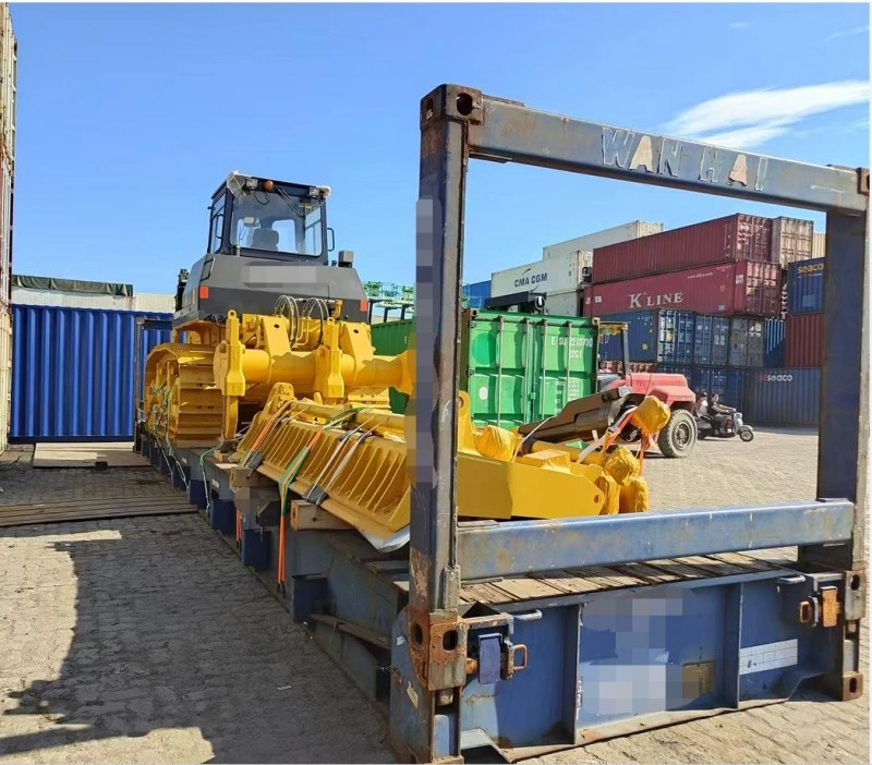 A large yellow bulldozer is securely positioned on a blue flat rack container in a shipping yard, surrounded by stacked standard containers, illustrating heavy equipment transport planning.