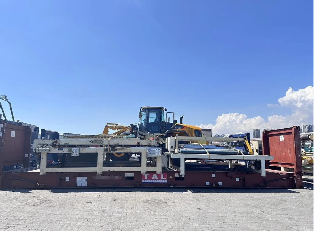 A yellow front-end loader mounted on a white steel support frame atop a red flat rack container, positioned outdoors at a port or industrial yard with urban skyline visible in background.