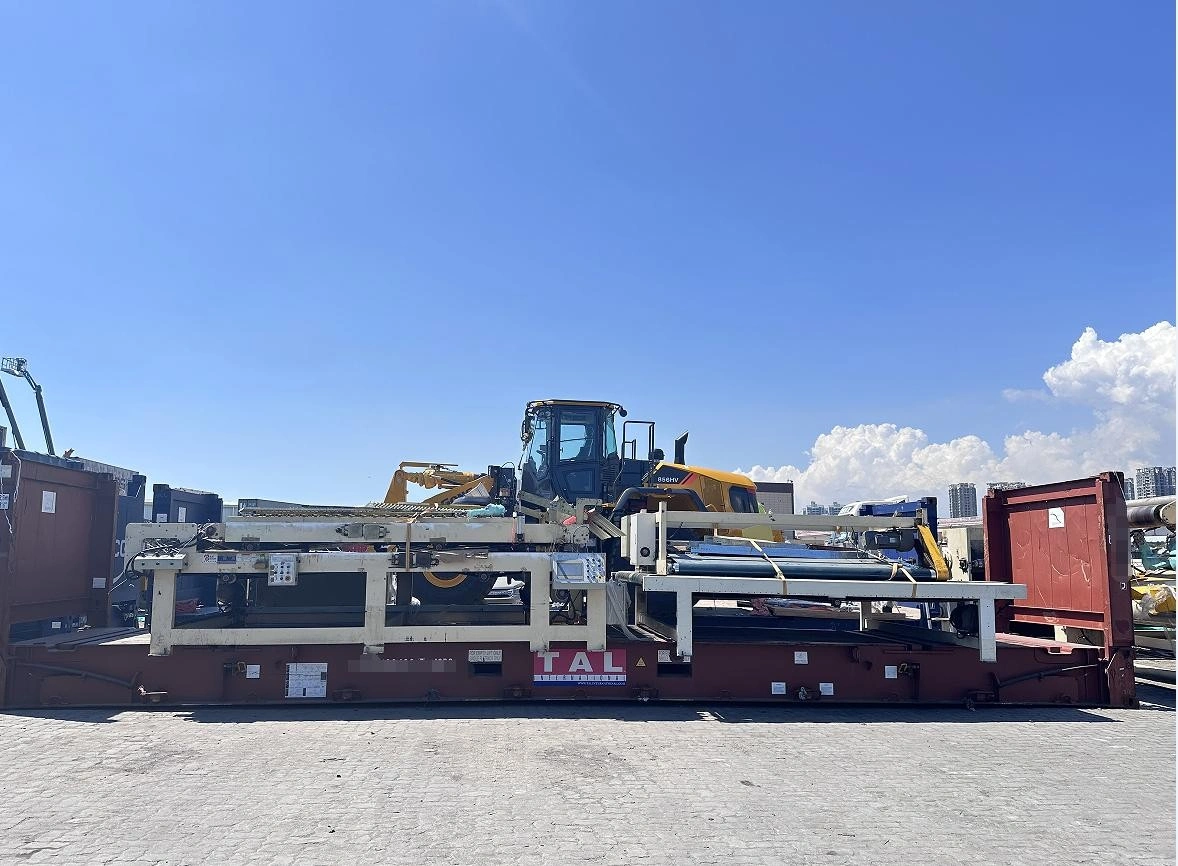A yellow front-end loader mounted on a white steel support frame atop a red flat rack container, positioned outdoors at a port or industrial yard with urban skyline visible in background.