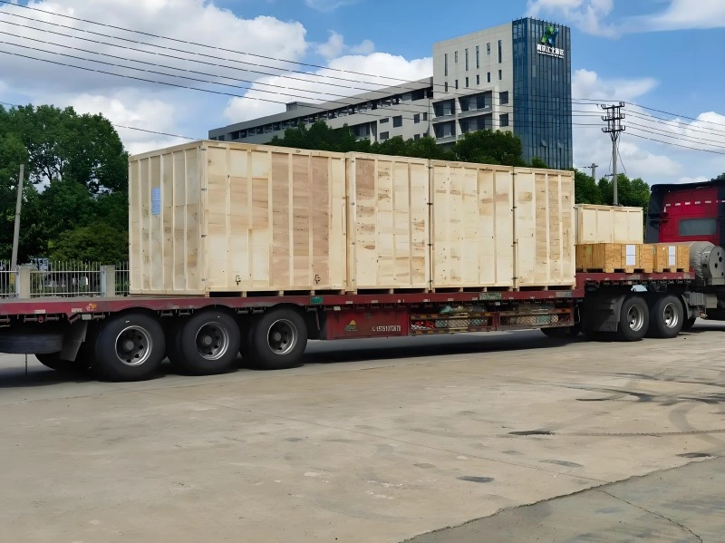 A red flatbed semi-trailer carrying multiple large, reinforced wooden export crates under a blue sky, demonstrating secure road transport solutions for oversized industrial equipment.