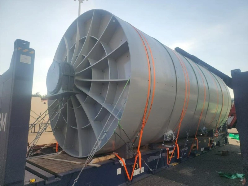 A side view of a large, gray industrial fan on a flat rack container, showcasing the intricate network of orange lashing straps and steel cables used to secure the heavy, cylindrical cargo against movement.