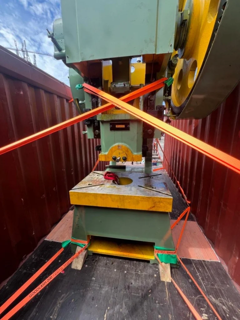 Close-up view of a large green and yellow industrial press machine secured inside a shipping container with bright orange cross-straps and wooden blocking.