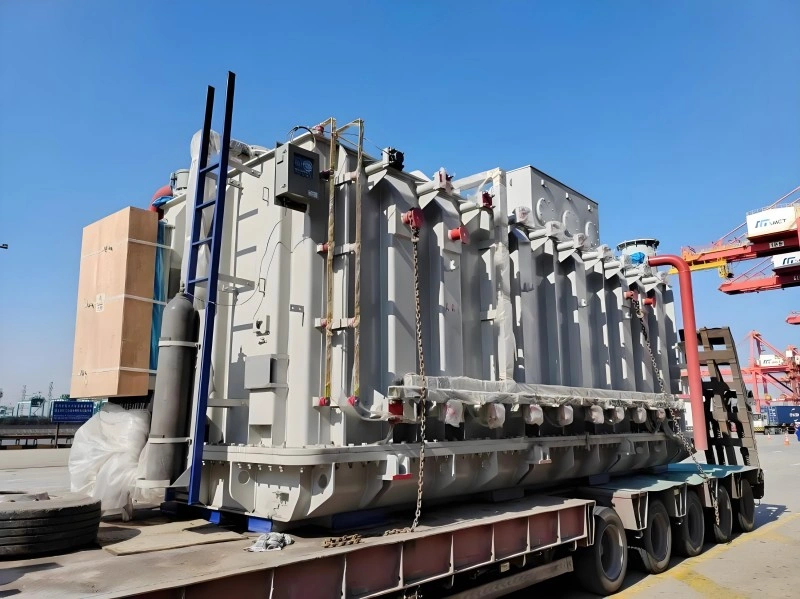 A massive gray power transformer secured with chains and straps rests on a red multi-axle flatbed trailer at a port terminal, with cranes and shipping containers visible in the background, demonstrating heavy equipment handling for grid infrastructure projects.