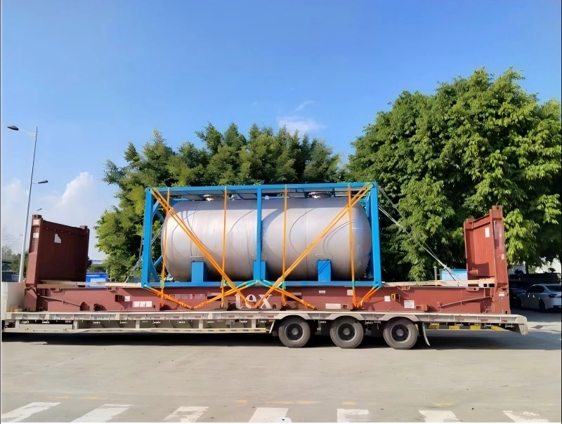 A close-up side view of a large, cylindrical vessel in a blue steel frame on a flatbed trailer. The vessel is secured with multiple orange straps, and the trailer is on a paved road with trees in the background. The text Bentlee is visible on the vessel.