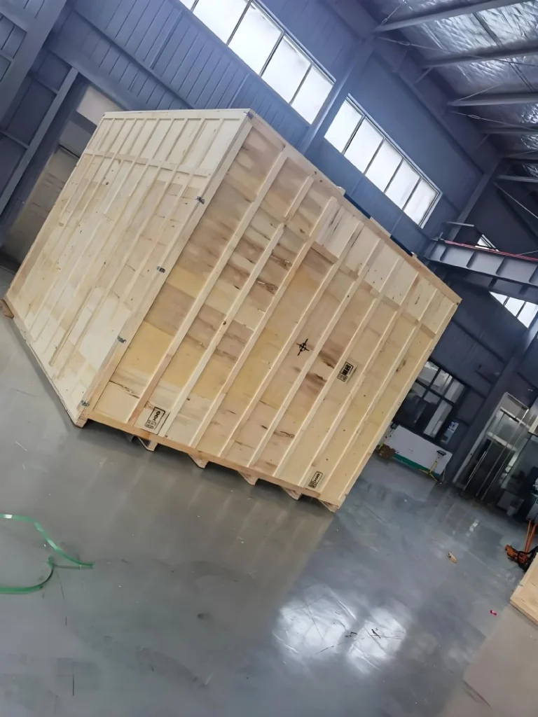 A large, reinforced wooden crate with visible structural framing and corner protectors, positioned inside a warehouse, designed for secure transport of heavy industrial equipment.
