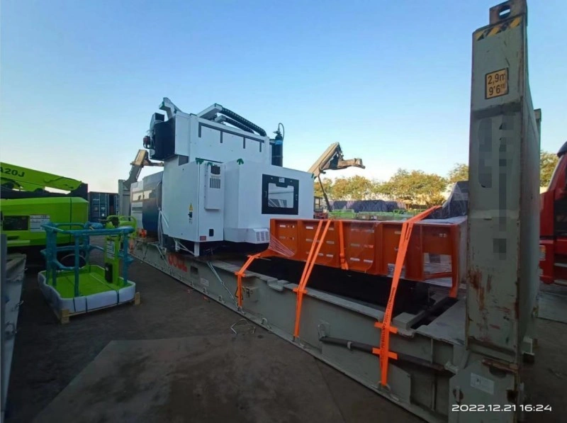 A large industrial machine being carefully positioned onto a flat rack container using lifting equipment, with safety personnel overseeing the operation in an outdoor logistics area.