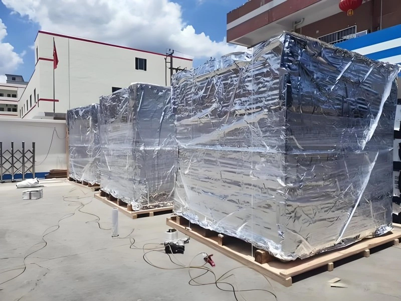 Blue industrial equipment fully enclosed in reflective aluminum foil vacuum bag on wooden skid, designed to prevent rust and salt spray damage during sea transport.