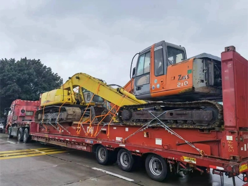 Two heavy excavators, one orange ZAXS 210 and one yellow model, are tightly lashed with orange straps onto a red lowbed trailer in an industrial yard, demonstrating secure transport methods for oversized construction equipment.