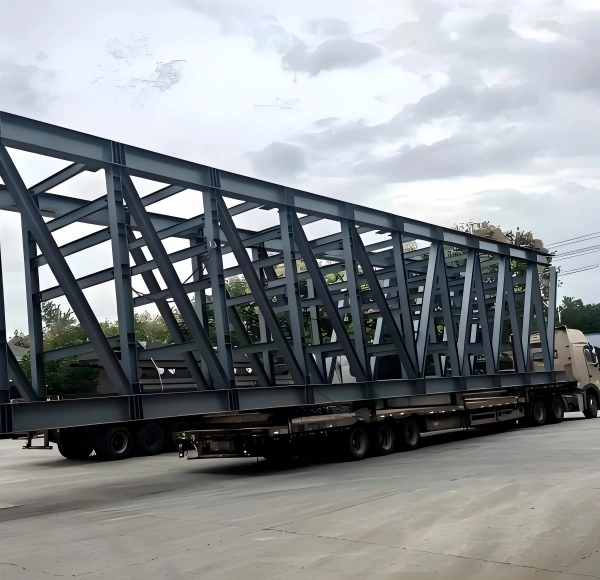 Specialized low-bed trailer carrying a massive grey steel bridge section, highlighting weight distribution and stability for heavy structural steel transport.