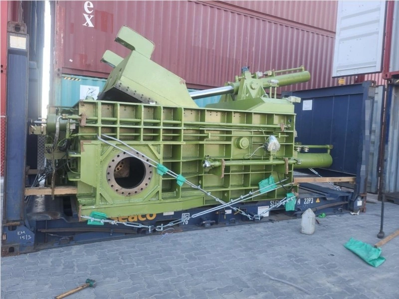 A large green industrial hydraulic press machine, secured with metal chains and wooden supports, is loaded onto a flat rack container in a port yard, ready for international transport.