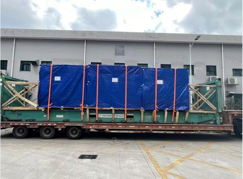 A long flatbed trailer carrying a large piece of industrial machinery wrapped in blue tarpaulin and secured with orange straps, parked outside a factory building under overcast skies.