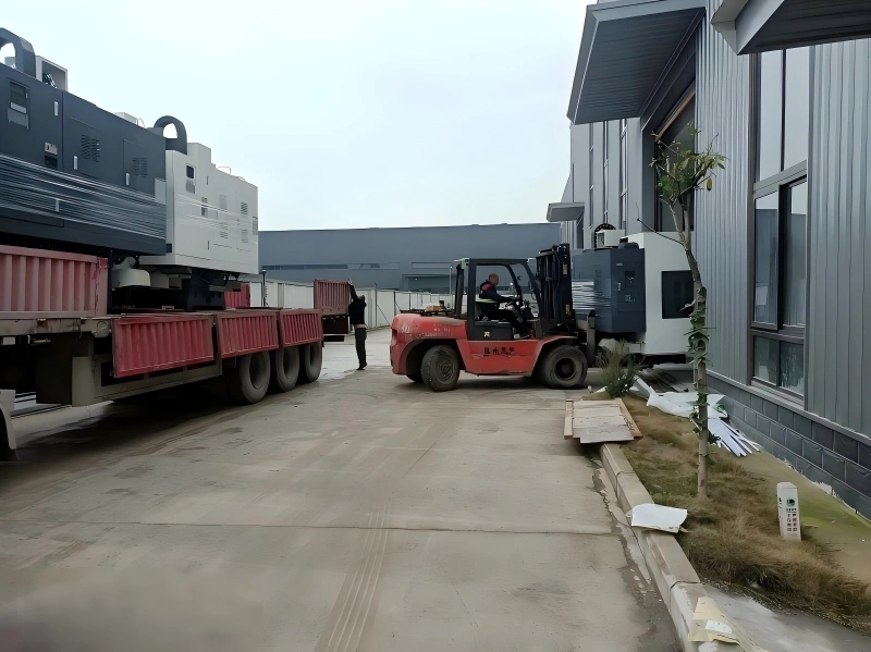 Workers using a forklift to load heavy manufacturing machinery onto a flatbed truck outside a factory building for safe transportation to a new site.