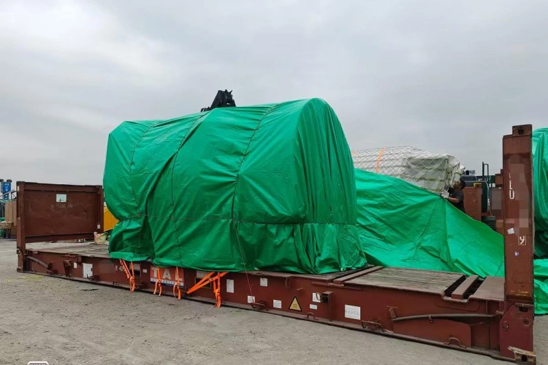 A large rectangular industrial machine, fully enclosed in a green waterproof tarpaulin and reinforced with orange tension straps forming diagonal cross-bracing, is mounted on a red flat rack container at an outdoor logistics yard under overcast skies.