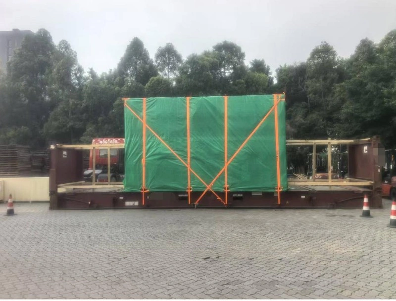 A large rectangular industrial machine, fully enclosed in a green waterproof tarpaulin and reinforced with orange tension straps forming diagonal cross-bracing, is mounted on a red flat rack container at an outdoor logistics yard under overcast skies.