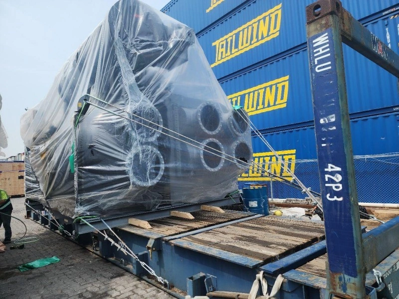 A large piece of industrial machinery wrapped in clear protective plastic is secured on a blue flat rack container using multiple steel wire ropes and wooden dunnage. The cargo is positioned in a shipping yard with stacked blue containers in the background.