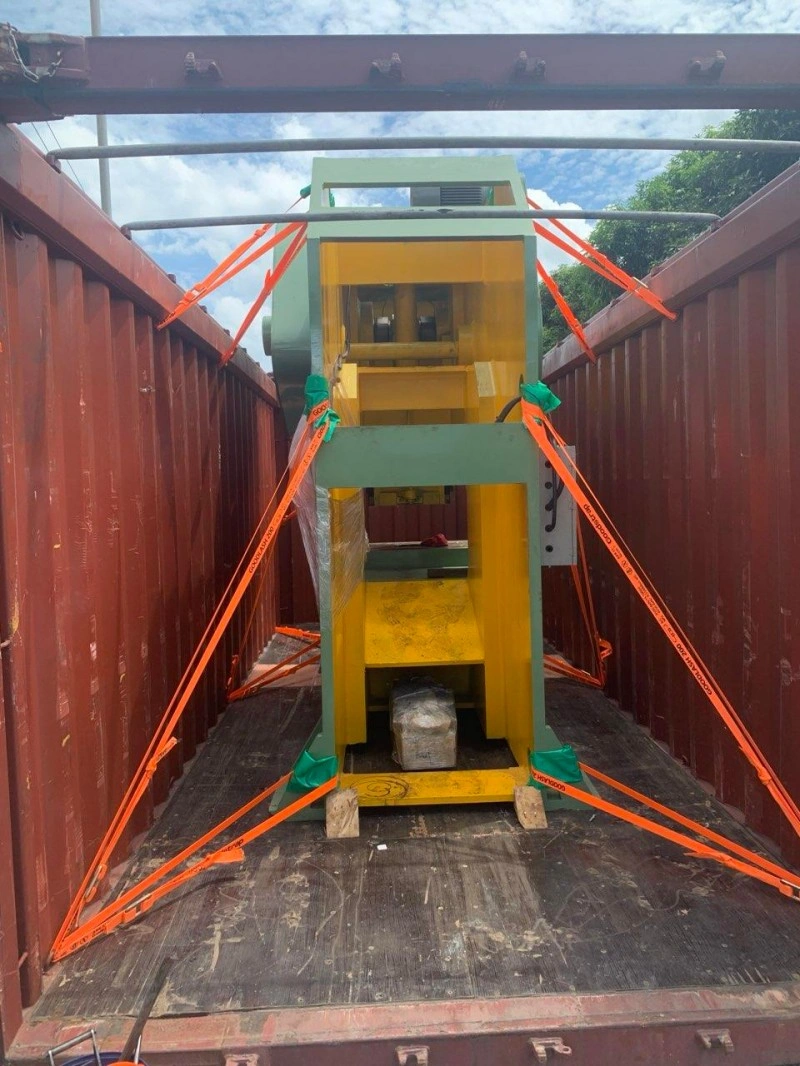 A massive grey industrial hopper with multiple chutes is being lifted by an overhead crane in a factory workshop, positioned above a flatbed trailer next to a shipping container.