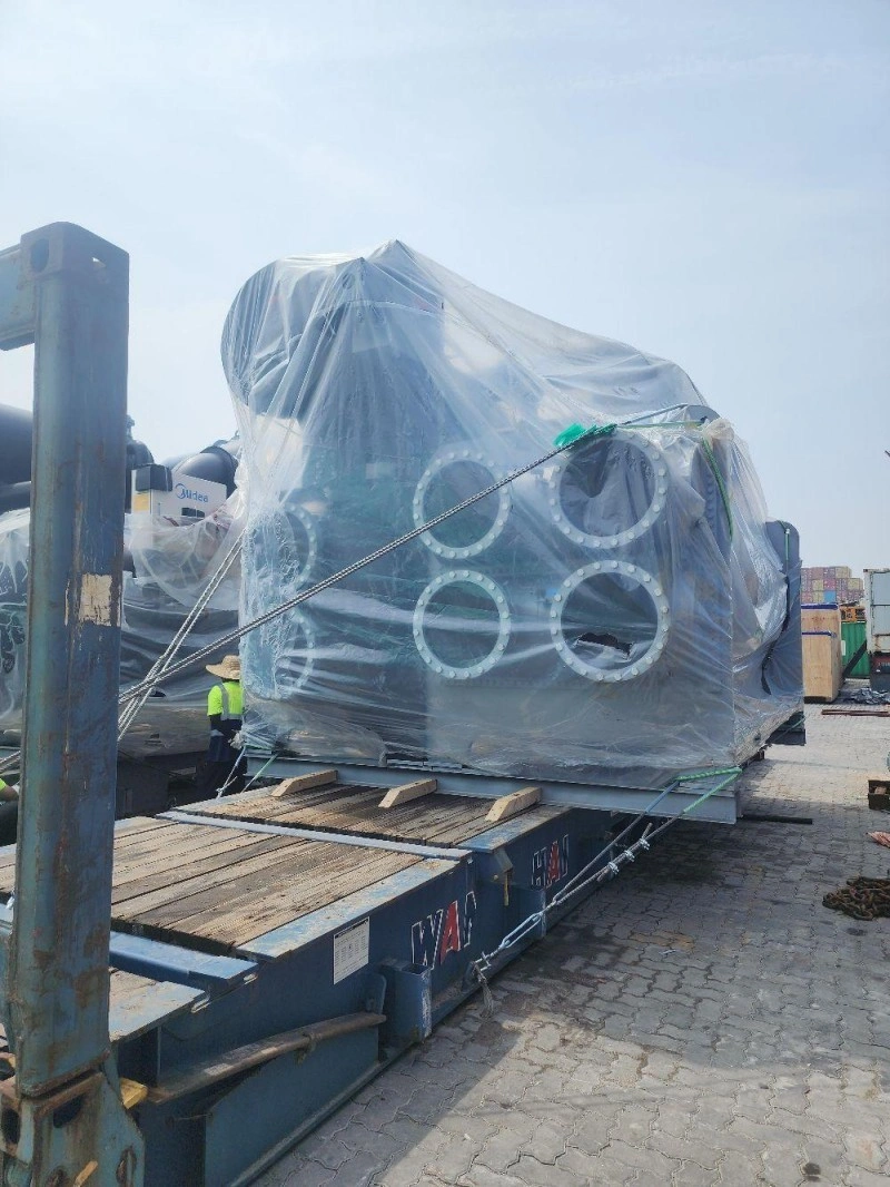 A massive piece of industrial equipment, fully encased in transparent shrink wrap and fastened with tensioned steel straps, rests on a blue flat rack container in an outdoor logistics yard under clear skies.