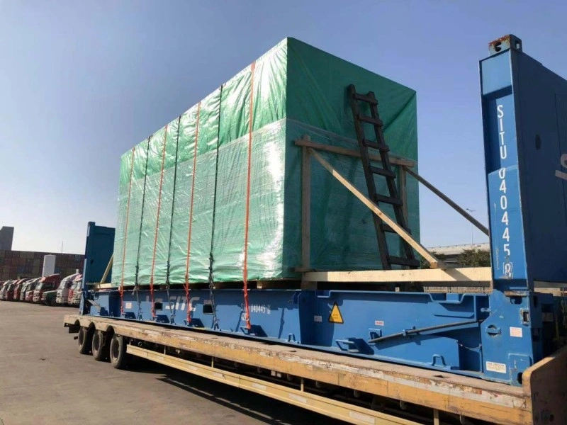 A large, irregularly shaped piece of industrial machinery, wrapped in clear protective film and black tarp, is loaded onto a red flat rack container. Multiple bright orange heavy-duty lashing straps are used to secure the cargo to the container's deck, with workers visible on top making adjustments in an outdoor logistics yard.