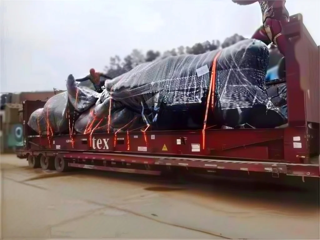A large, irregularly shaped piece of industrial machinery, wrapped in clear protective film and black tarp, is loaded onto a red flat rack container. Multiple bright orange heavy-duty lashing straps are used to secure the cargo to the container's deck, with workers visible on top making adjustments in an outdoor logistics yard.