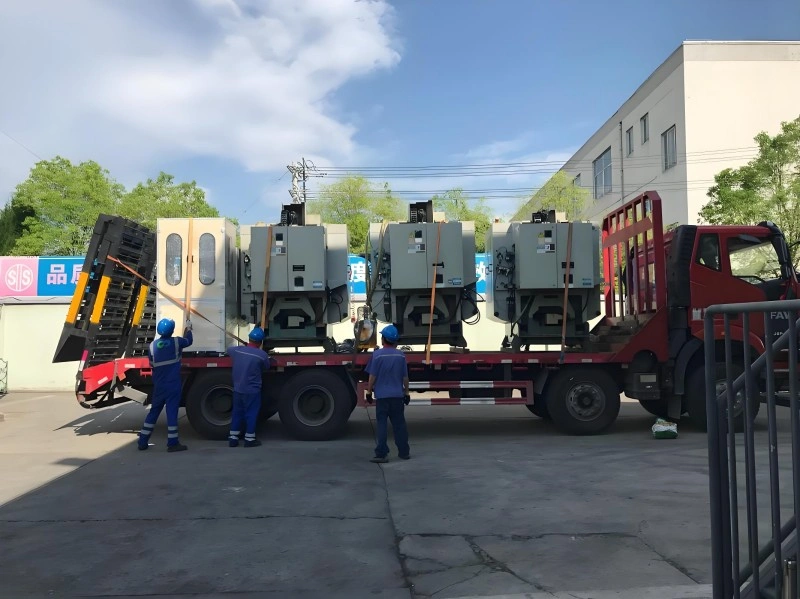 Workers in blue uniforms securing large grey industrial machines onto a red flatbed truck at a factory yard.