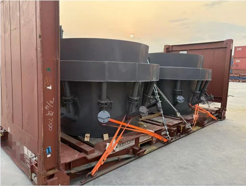 Two large black cylindrical pressure vessels mounted side-by-side on a red flat rack container, secured with orange tension straps and metal turnbuckles; background shows stacked shipping containers under twilight sky.