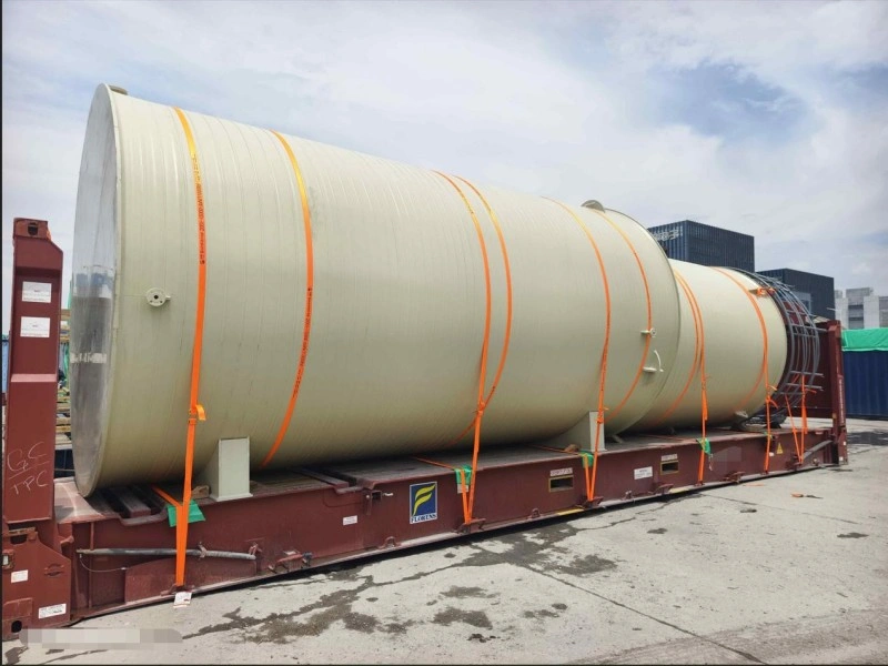 A large, smooth-surfaced industrial tank, secured with multiple orange straps, rests on a blue Seaco-branded flat rack container at a port terminal under a partly cloudy sky, with other containers and yellow handling equipment nearby.