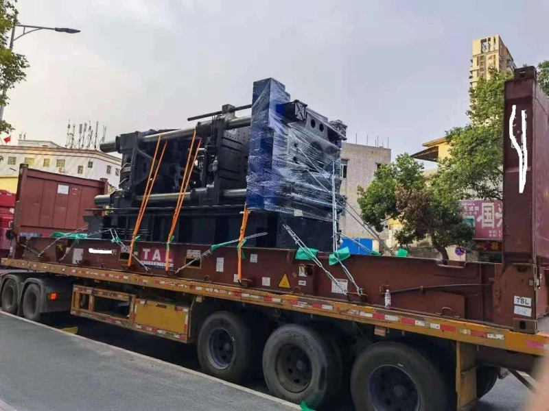 A large black industrial injection molding machine, partially wrapped in blue protective film and secured with orange lashing straps, is loaded onto a flat rack trailer marked with “TAL” branding, ready for road transport to port.