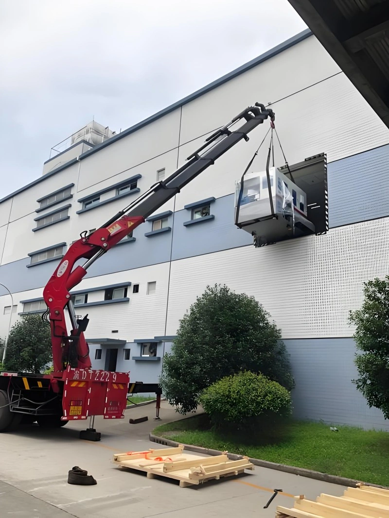 A red mobile crane lifting a packaged industrial unit through a second-floor window or opening of a factory building for installation.