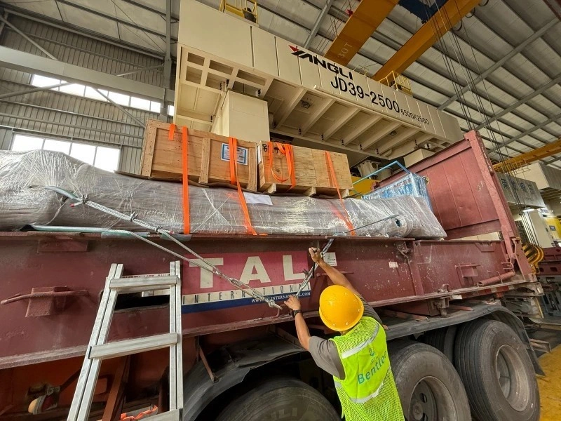 Workers securing heavy industrial equipment loaded onto a flatbed truck using an overhead crane, ensuring safe transport.