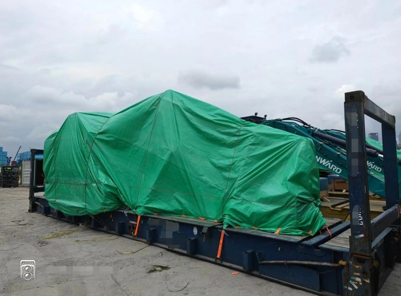 Large industrial machinery wrapped in green protective tarpaulin and secured with orange lashing straps on a specialized flatbed trailer, demonstrating safe transport protocols for oversized equipment during factory moves.