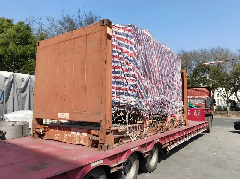 Large industrial equipment wrapped in red, white, and blue striped tarpaulin and secured with cargo netting on a flatbed trailer next to an open-top shipping container frame for export logistics.