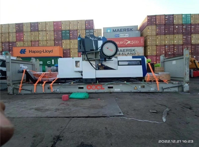A large industrial machine is loaded onto an OOCL flat rack container, secured with orange lashing straps, with stacked shipping containers from Hapag-Lloyd, Maersk, and Hamburg Süd in the background