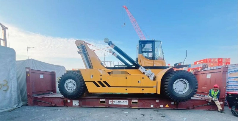 A yellow heavy-duty reach stacker is securely positioned on a red flat rack container with visible lashing points, under clear sky at a logistics yard, demonstrating specialized transport for oversized construction equipment.