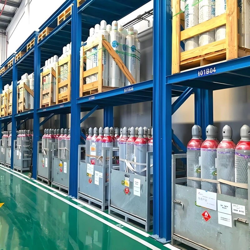Interior view of a modern chemical warehouse with tall blue and orange pallet racking. The shelves hold numerous blue plastic drums on upper levels and large white Intermediate Bulk Containers (IBCs) with hazard labels on lower levels, all on a polished green epoxy floor.
