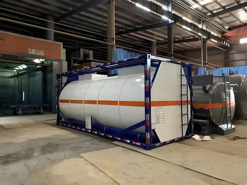 A blue-framed ISO tank container with white cylindrical tanks and orange hazard stripes is positioned inside a large industrial manufacturing facility. The container is resting on a concrete floor with factory equipment and structural elements visible in the background.